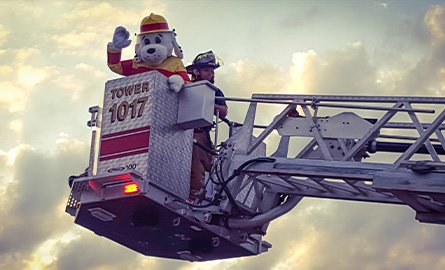 Photo of Sparky and a firefighter standing in the bucket of an erected firetruck ladder