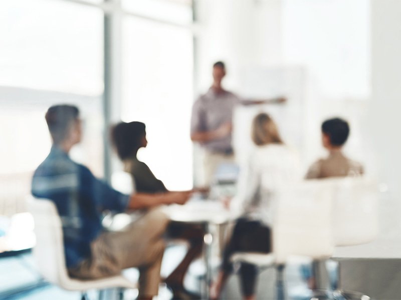 A group of people sitting around a desk while one person gives a presentaion.