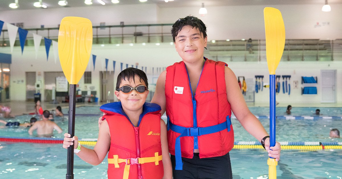 Two people wearing life jackets holding paddles inside of the BWG Leisure Centre Pool area.
