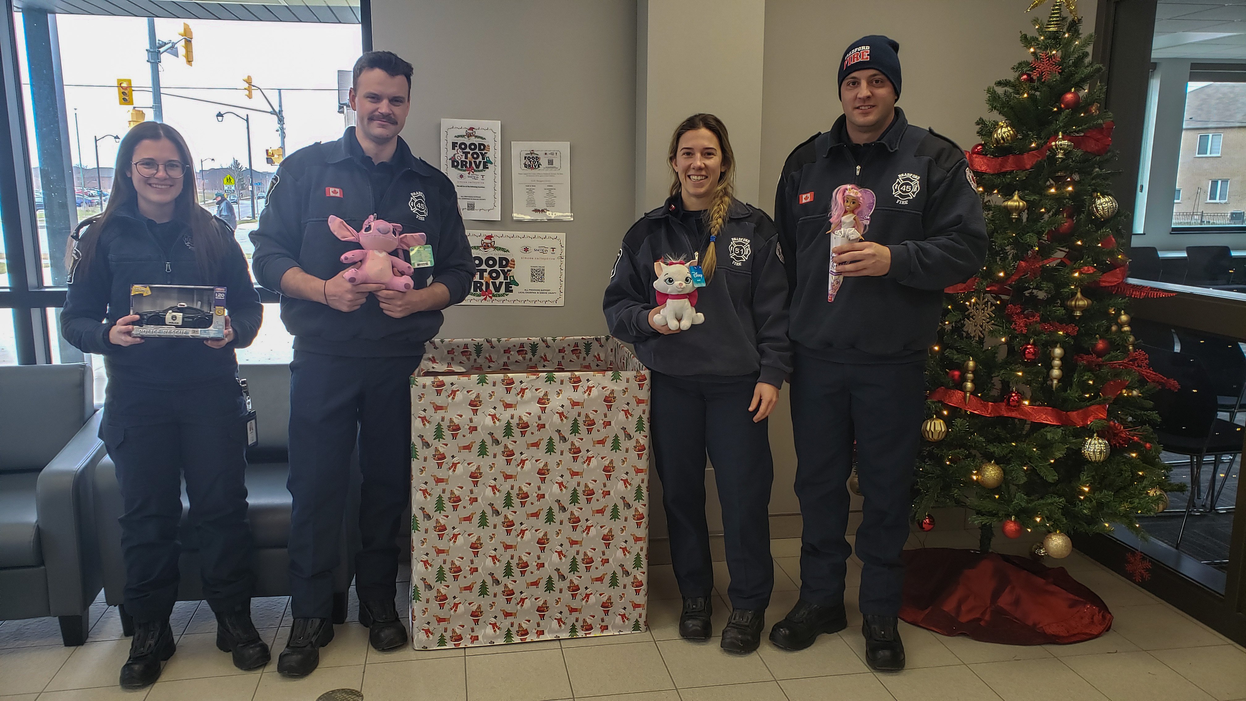 Four BWG Fire and Emergency Services workers smiling holding toys inside of the BWG Fire Hall standing beside a Christmas tree and toy box.